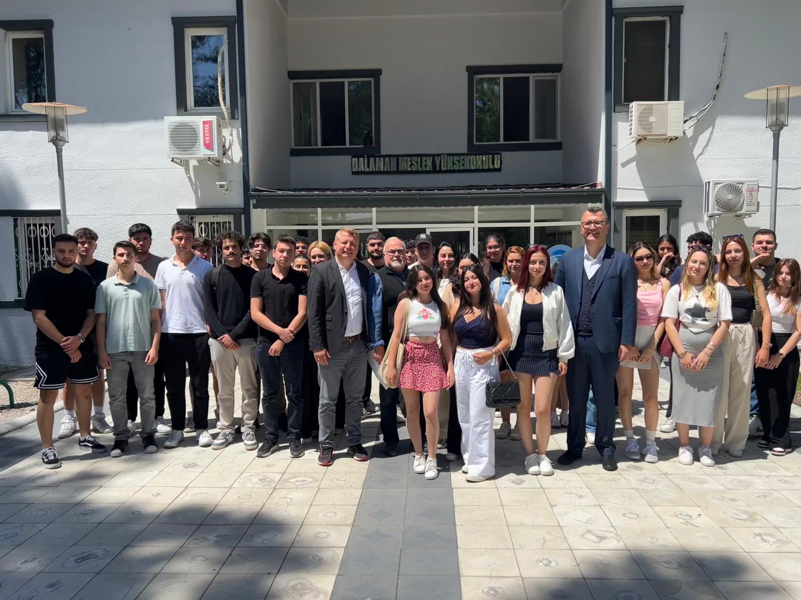 Dalaman Meslek Yüksekokulu binasının önünde, çoğunluğu gençlerden oluşan yaklaşık 35 kişilik bir grup toplanmış. Ortada takım elbise giymiş birkaç yetişkin erkek ve kadın yer alıyor, muhtemelen öğretim görevlileri ya da yöneticiler. Etraflarında ise öğrenciler rahat kıyafetlerle ayakta poz veriyor. Fotoğraf güneşli bir günde, binanın giriş kapısı önünde çekilmiş.